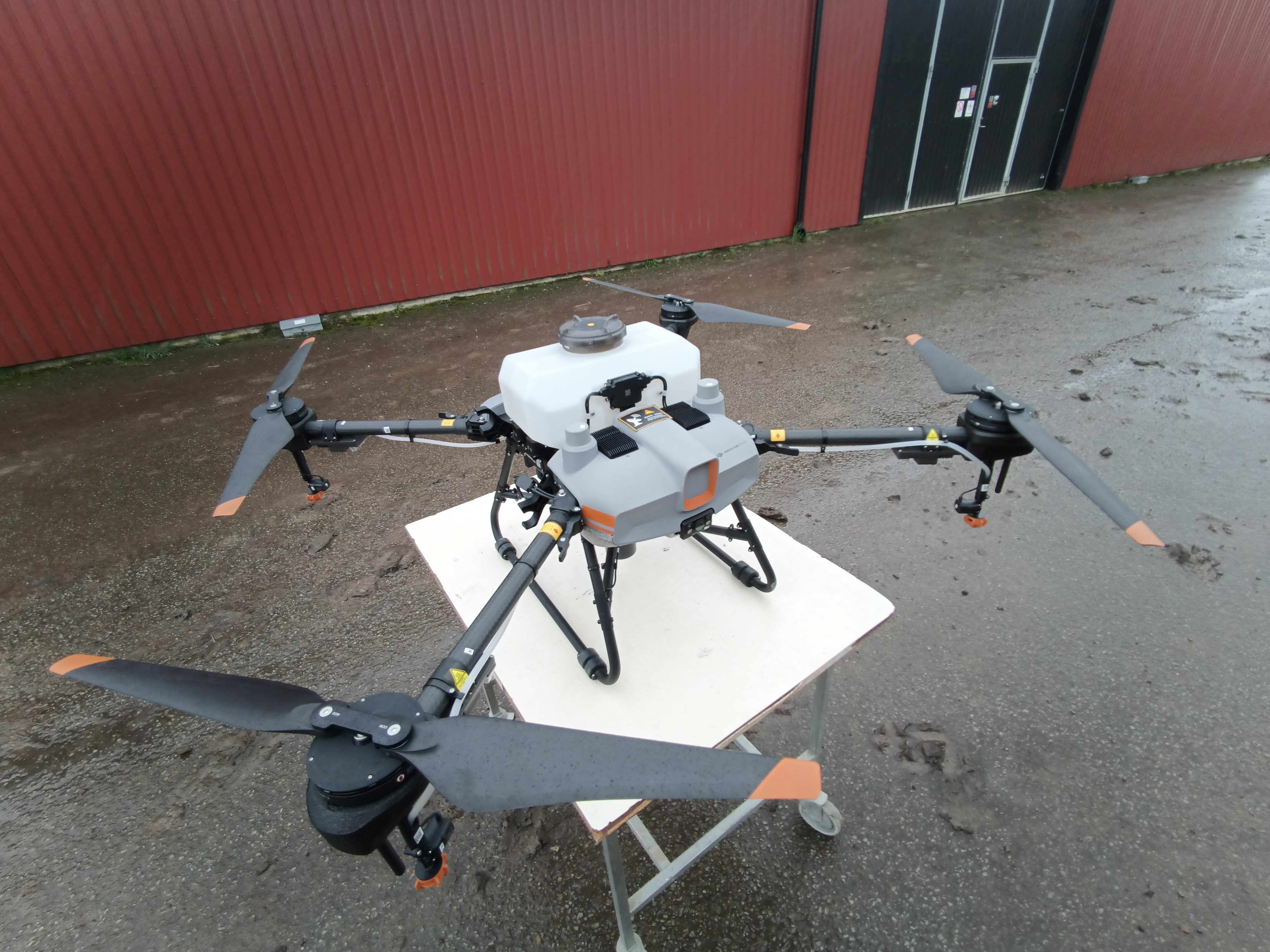 A photo of a drone sitting on a little cart in front of a red building. The drone has four propellers on arms reaching away from the body of the drone where the there is the container that could be filled with fertilizer.